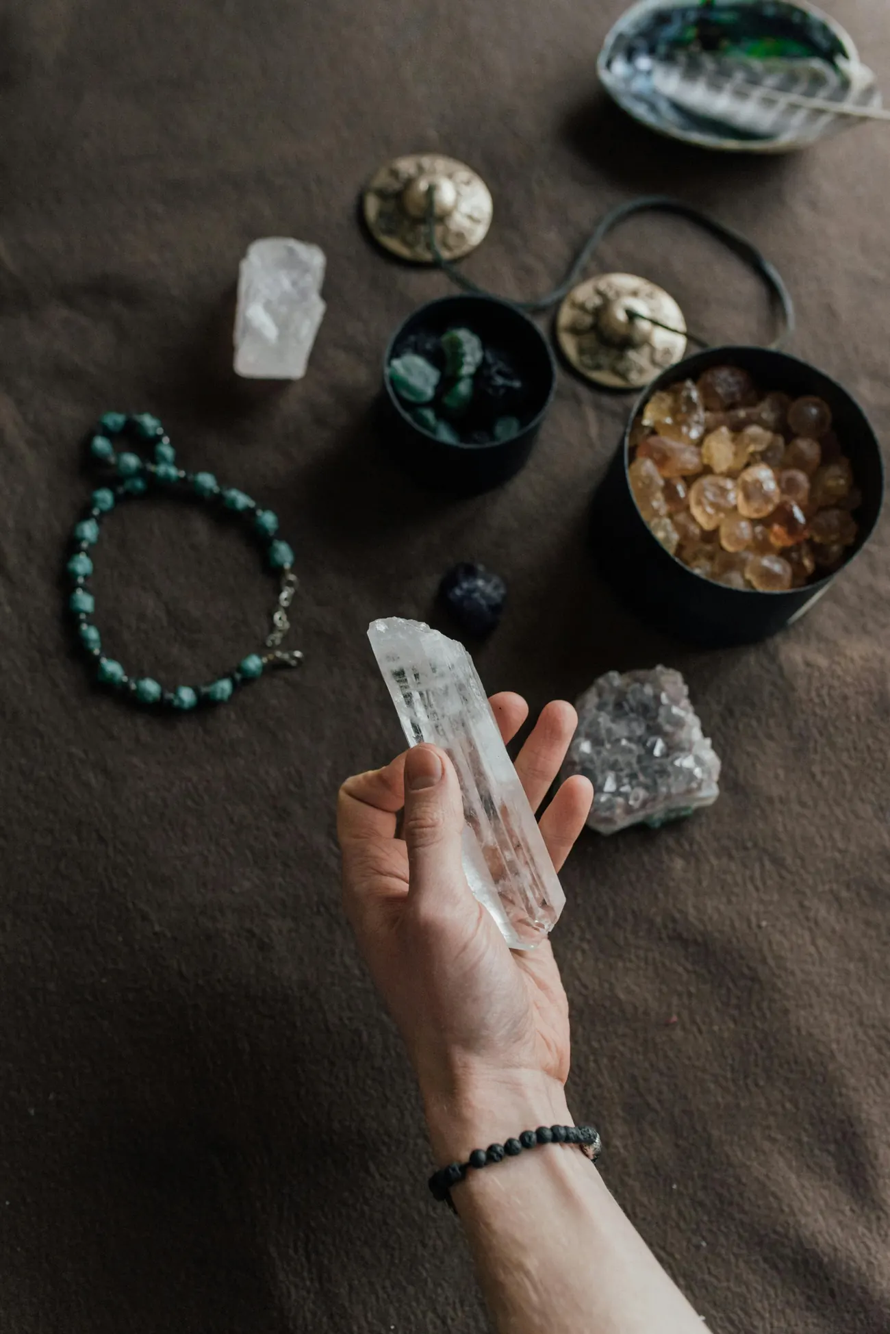 A hand holding a clear quartz crystal above bowls of tumbled stones