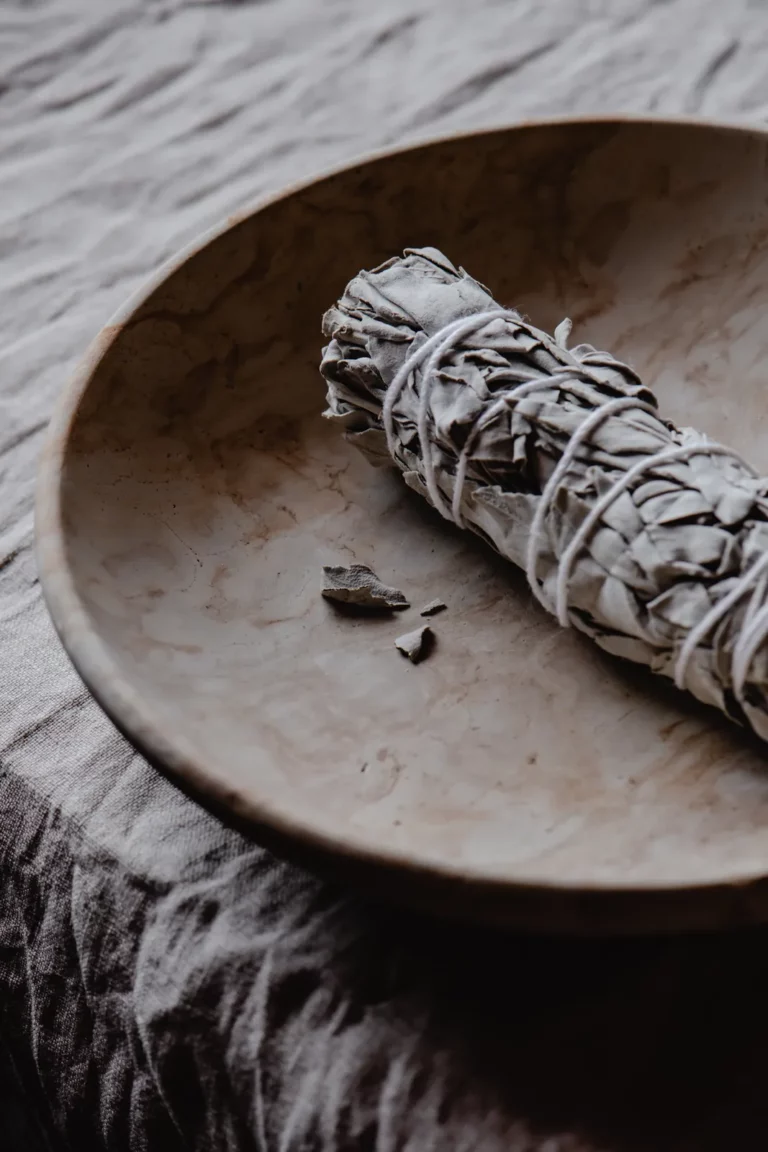 A bundle of white sage resting in a pale stone bowl on linen